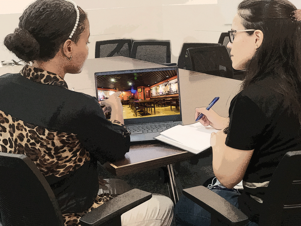 A imagem mostra Roberta e Laura conversando no escritório da SI Construções sobre o projeto, Laura está apresentando em um notebook a perspectiva que criou do ambiente com as modificações solicitadas pelo cliente.